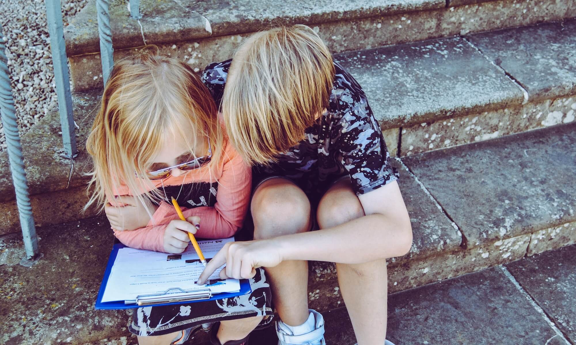 Two children sitting on steps working together on a document on a clipboard